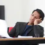 Ondernemer aan bureau omringd door papieren, voelt zich overweldigd en uitgeput door werkdruk