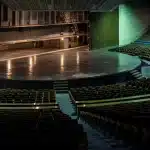 Theatercriticus zittend in een theaterzaal met notitieboekje, kijkend naar een verlicht podium